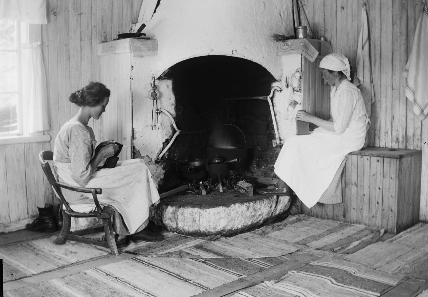 Två kvinnor som handarbetar framför vedspis. Golvet täckt av trasmattor.